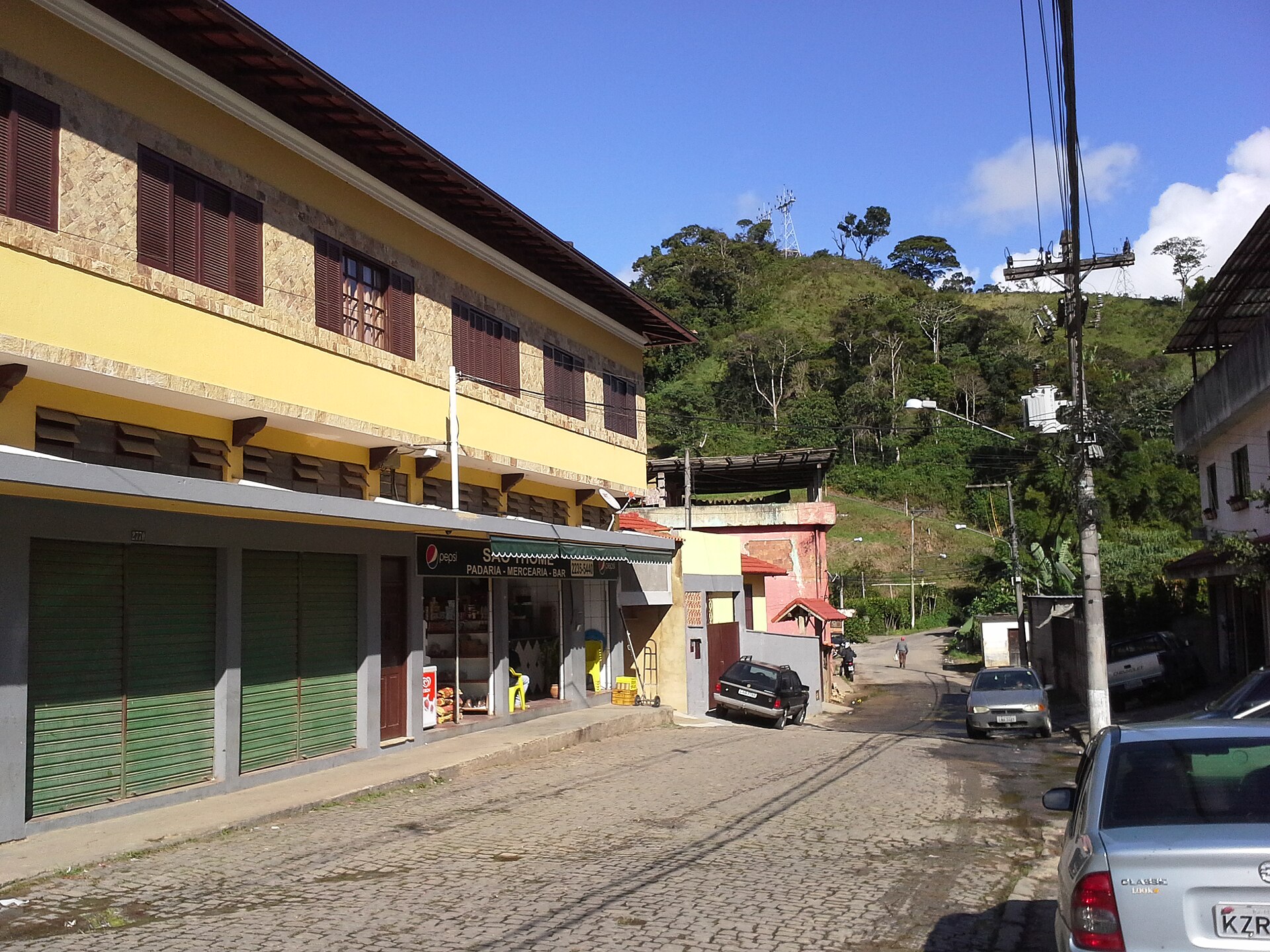Padaria de bairro em uma rua de Petrópolis, com morros verdes ao fundo.