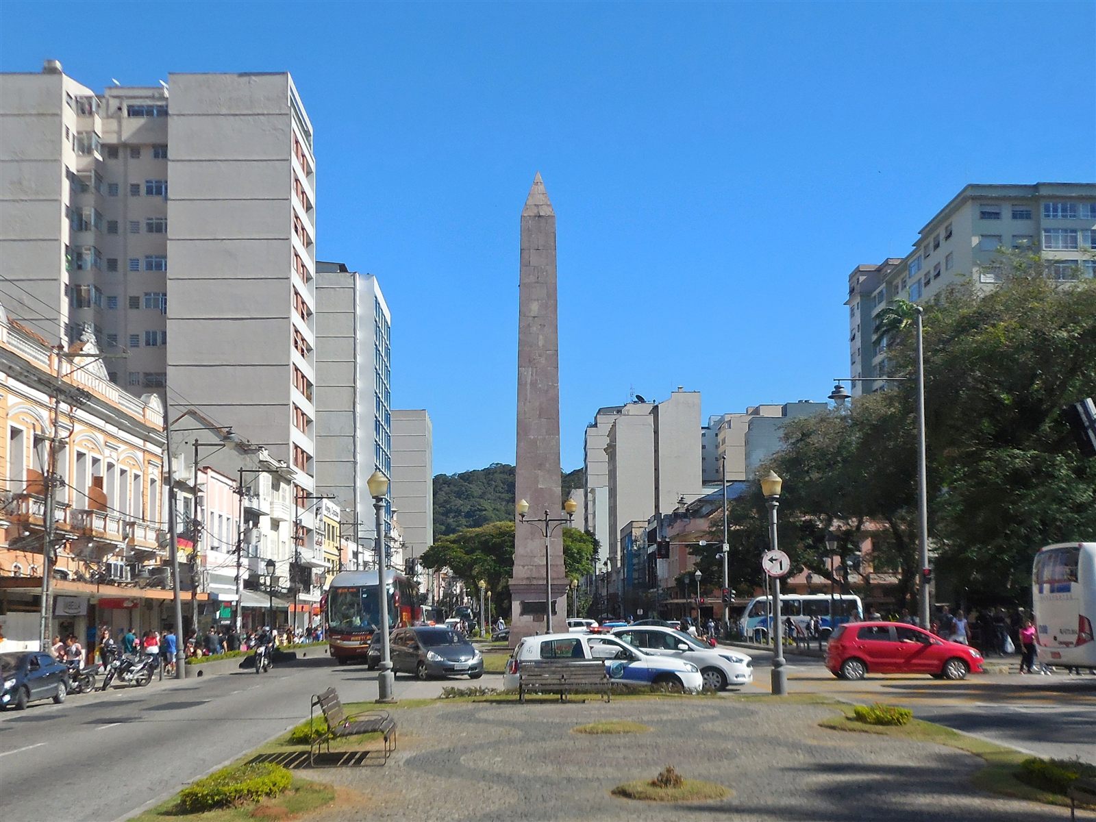Praça do Obelisco no Centro de Petrópolis, com o monumento, prédios, comércio, veículos e pedestres.